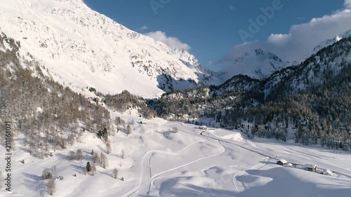 Aerial flight in a snowy winter valley in the swiss alps towars peaks over trees in sunset light.