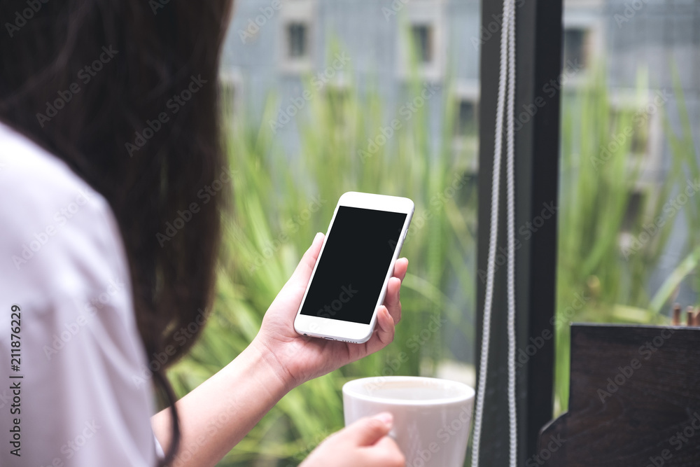 Mockup image of woman holding white mobile phone with blank black screen while drinking coffee in cafe