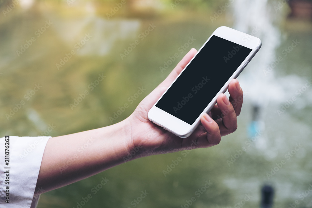 Mockup image of a woman's hand holding white smart phone with blank black desktop screen with blur green nature background