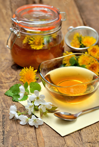 Close-up of dandelion honey, spring flowers, spoon, dandelion head around, full jar in background - vertical photo