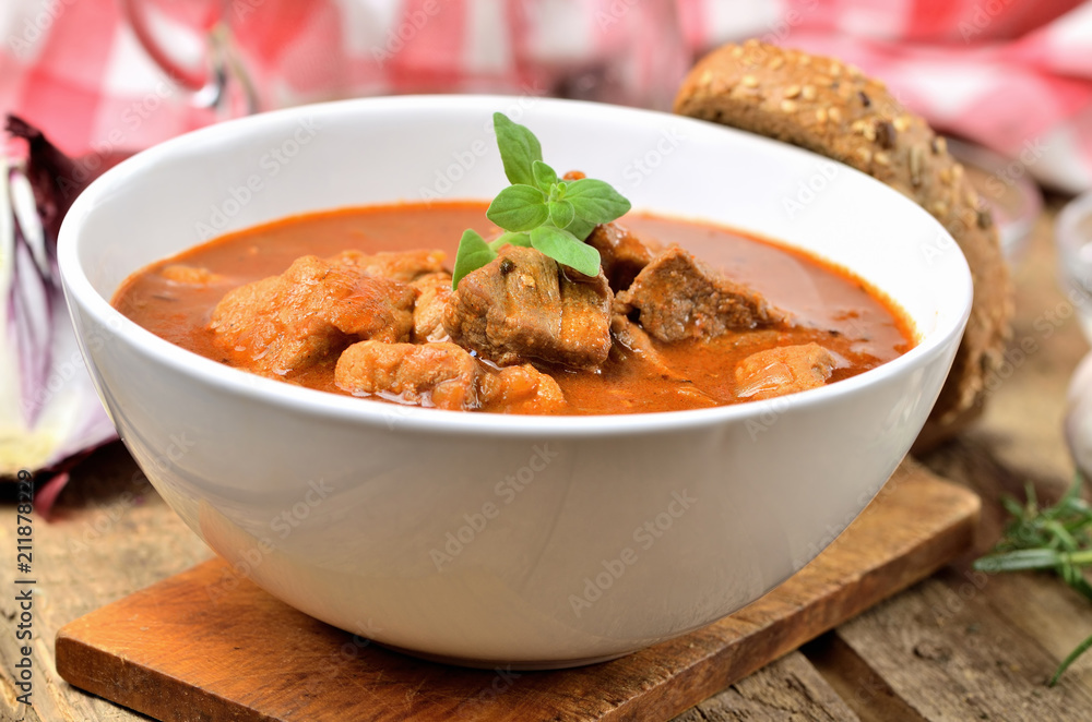 Close-up of pork goulash with pieces of meat in a bowl, oregano pepper, onion, red checkered tablecloth in the background Close-up of pork goulash with pieces of meat in a bowl, oregano pepper, onion, red checkered tablecloth in the background