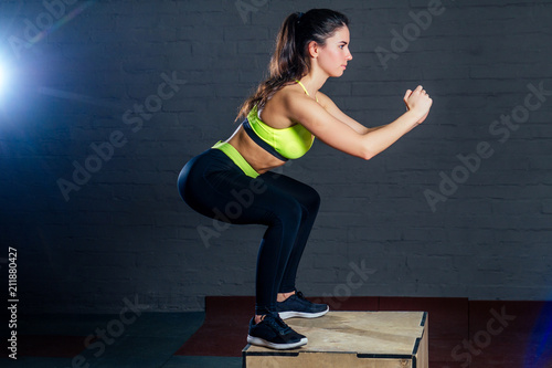 a young woman in a green sports bra and black pants jumps on a box against a backdrop of black bricks