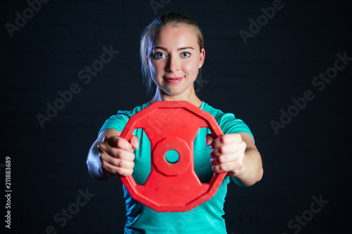 young woman in turquoise t-shirt and black pants raises pancake from the bar on a black background in the gym