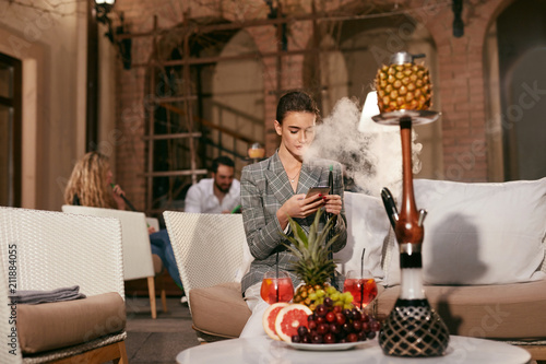Woman With Phone Smoking Fruit Hookah In Shisha Bar photo