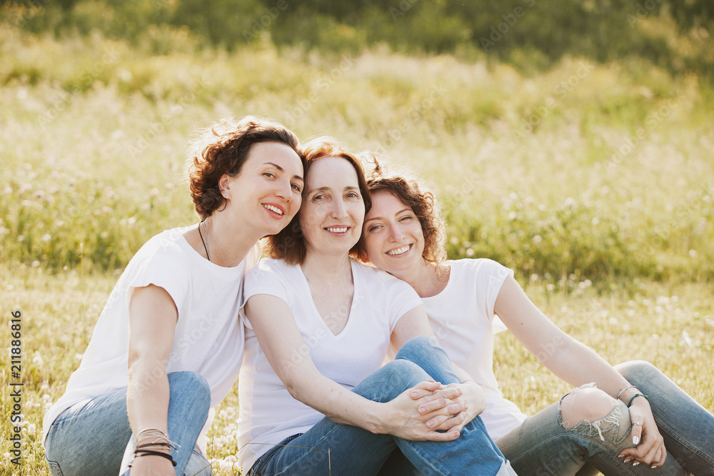 Continuity of generations. Portrait of three beautiful women - mother and adult daughters sitting on the field wearing the same clothes white t-shirts and blue jeans on sunny summer day Continuity of generations. Portrait of three beautiful women - mother and adult daughters sitting on the field wearing the same clothes white t-shirts and blue jeans on sunny summer day