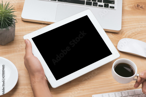 Mock up image of a hand holding black tablet pc with white blank screen and coffee cup on wooden table background