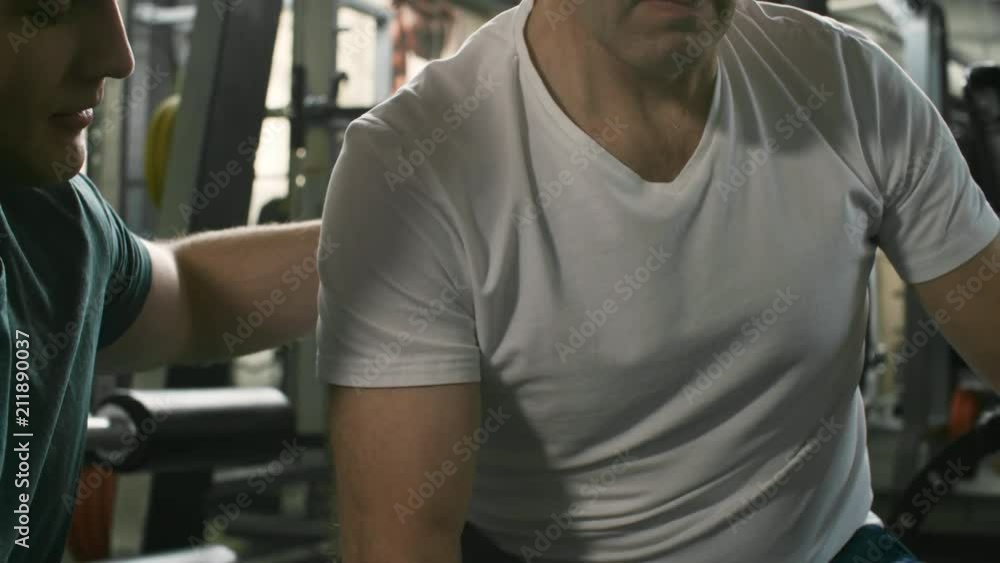 Tilt up shot of middle-aged sportsman doing exercises for biceps using dumbbell when working out at the gym under guidance of coach Tilt up shot of middle-aged sportsman doing exercises for biceps using dumbbell when working out at the gym under guidance of coach