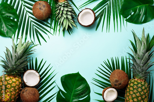Exotic pineapples, ripe coconuts, tropical palm and green monstera leaves on blue background with copyspace for your text. Creative layout. Summer concept. Flat lay, top view