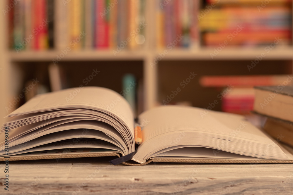 Notepad / book is on a wooden table. orange simple pencil. background, bookcase with colored books Notepad / book is on a wooden table. orange simple pencil. background, bookcase with colored books