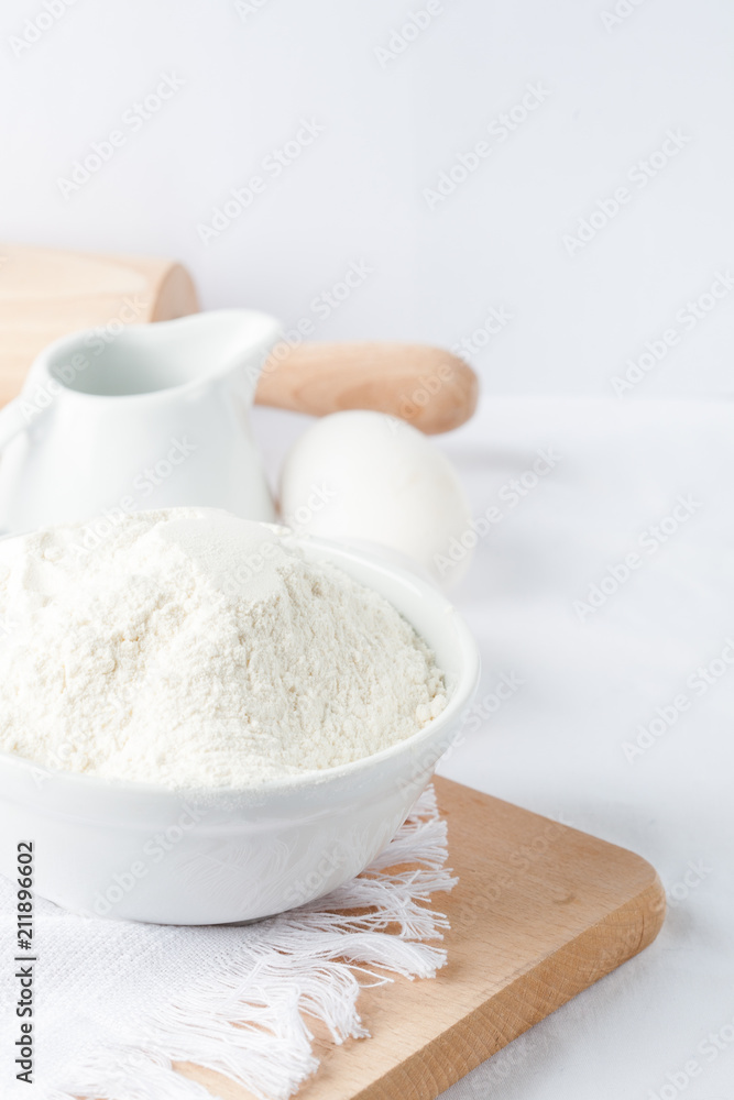  Baking ingredients. White background. Ingredients and tools to make a cake. Flour, milk, eggs, rolling pin, wooden spoons, jar. Copy space. Bakery. Baking concept