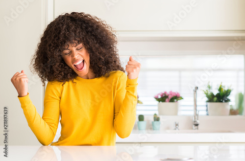 African american woman wearing yellow sweater at kitchen very happy and excited doing winner gesture with arms raised, smiling and screaming for success. Celebration concept.