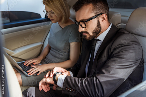handsome businessman checking time and assistant working with laptop in car