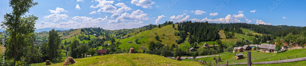 A charming panorama of mountainous green slopes on a summer day with rural houses scattered around the country with dry harvested hay on farms and white rare clouds floating on the horizon line.
