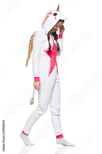 A side view of a young Asian woman wearing a unicorn kigurumi costume with a tail, gold wings and horn. The girl clad in pajamas smiling and looking down while walking against the white background. photo