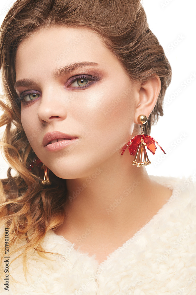Fototapeta premium Close up portrait of a young fair-haired lady with one sided hairstyle. The girl in an ivory shaggy coat and red chunky flower earrings, posing over the white background, looking at the camera.