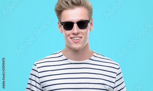 Young handsome blond man wearing sunglasess with a happy face standing and smiling with a confident smile showing teeth