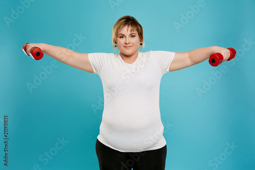 Three quarter isolated studio portrait of a beautiful chubby woman working out with dumbbells. The confident lady exercising over the blue background, looking at the camera. Body positive concept. photo