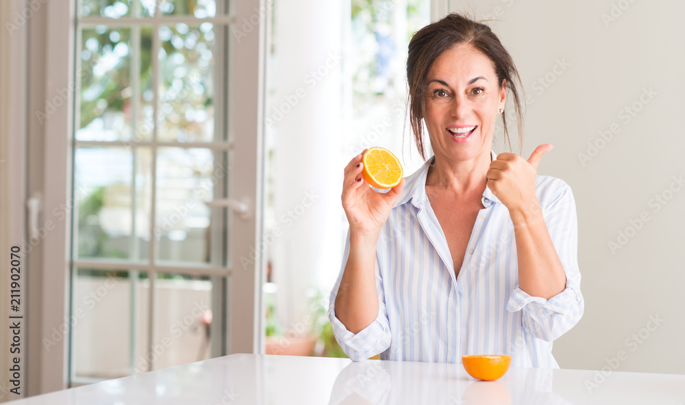 Fototapeta premium Middle aged woman holding orange fruit pointing with hand and finger up with happy face smiling