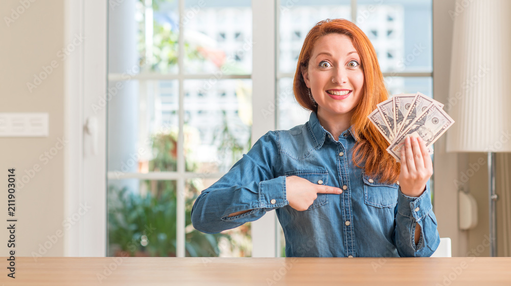 Redhead woman holding dollar bank notes at home with surprise face pointing finger to himself Redhead woman holding dollar bank notes at home with surprise face pointing finger to himself