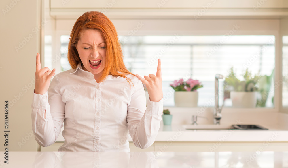 Redhead woman at kitchen shouting with crazy expression doing rock symbol with hands up. Music star. Heavy concept.