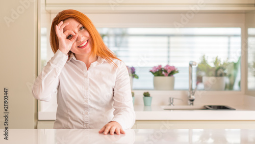 Redhead woman at kitchen doing ok gesture with hand smiling, eye looking through fingers with happy face.