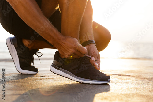 Cropped photo legs of sporty caucasian man in tracksuit and sneakers squatting on pier at seaside, and tying his shoelaces photo