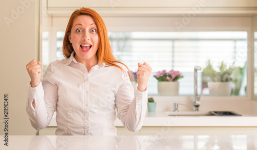 Redhead woman at kitchen celebrating surprised and amazed for success with arms raised and open eyes. Winner concept.