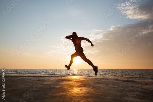 Silhouette of athletic guy 30s in shorts and t-shirt training and running along pier at seaside, and listening to music via wireless headphones during sunrise