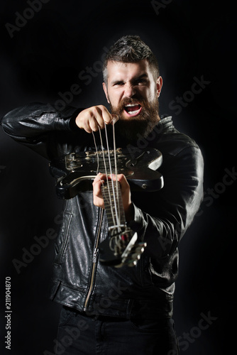 Bearded man with electric guitar. Guitarist in black leather jacket stands with bass guitar. Charismatic&stylish man with beard&mustache holds guitar pulls strings. Hobby, music, instrument education.