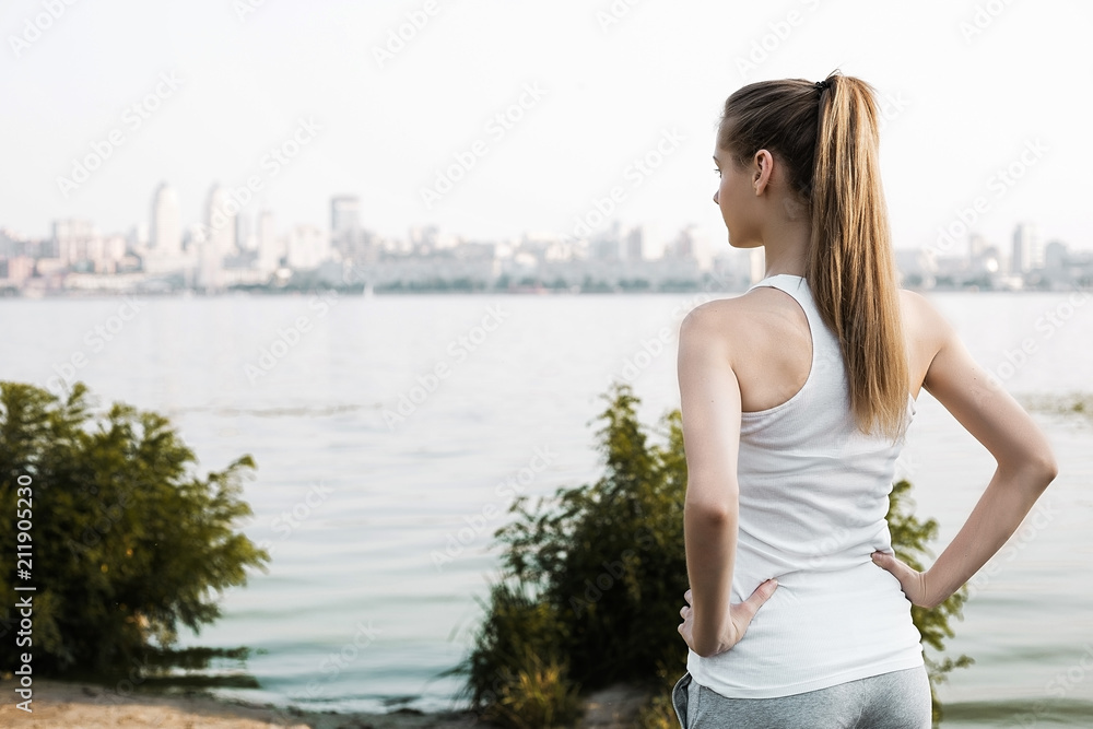 Pretty and young woman in sports clothes standing with her back to the camera and looking on the city behind river. Pretty and young woman in sports clothes standing with her back to the camera and looking on the city behind river.