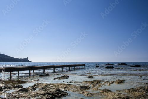 On the left side of the picture you can see the jetty. In the foreground flat rocks can be seen that look out of the water. In the background you can see a small piece of land, blue sea and blue sky. © ELKOKREATIV