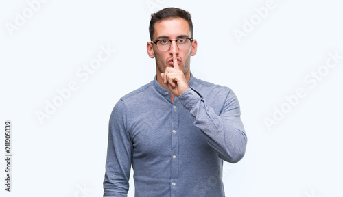 Handsome young elegant man wearing glasses asking to be quiet with finger on lips. Silence and secret concept.