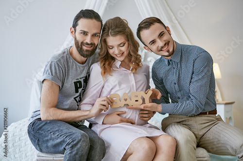 Gay couple becoming parents through surrogacy. The fathers-to-be sitting on the bed, a  surrogate mom between them, happy smiles on their faces. The men holding lettering BABY over the pregnant belly.