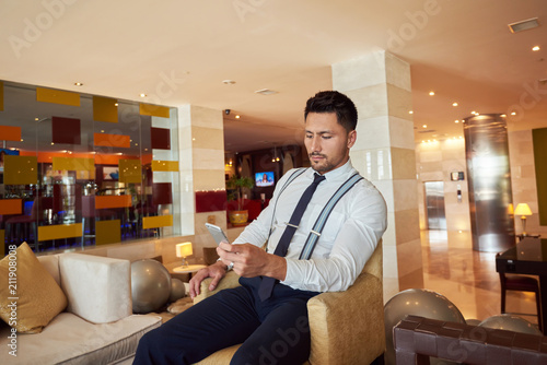 Man professional trader is reading good news on mobile phone from his client about purchase of securities, while is sitting in the lobby of an expensive hotel photo