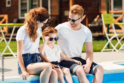 young family in white t-shirts and sunglasses sitting on poolside together