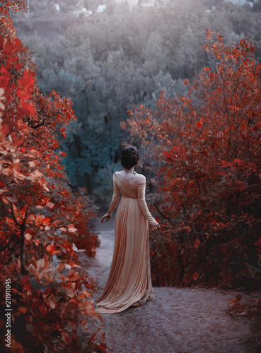 The princess in a yellow vintage dress in the Renaissance is walking along the picturesque autumn hills at dusk. Photo of a brunette girl with a back without a face. Artistic Photography.