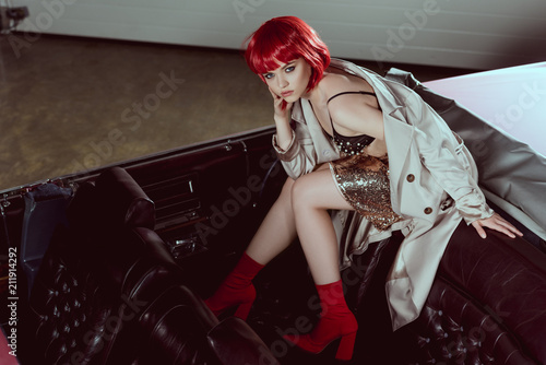 high angle view of stylish girl in trench coat looking at camera while sitting on classic car