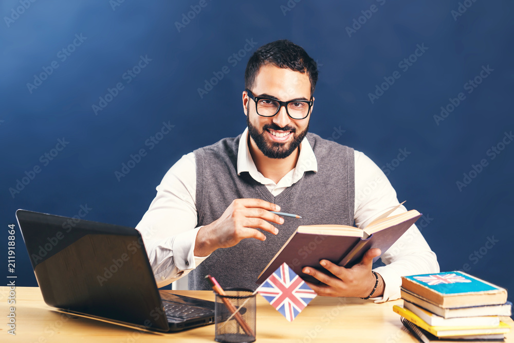 Black-haired bearded man learning english, wearing gray sweater vest and pristine white shirt, working hard at the office desk with laptop before black wall Black-haired bearded man learning english, wearing gray sweater vest and pristine white shirt, working hard at the office desk with laptop before black wall