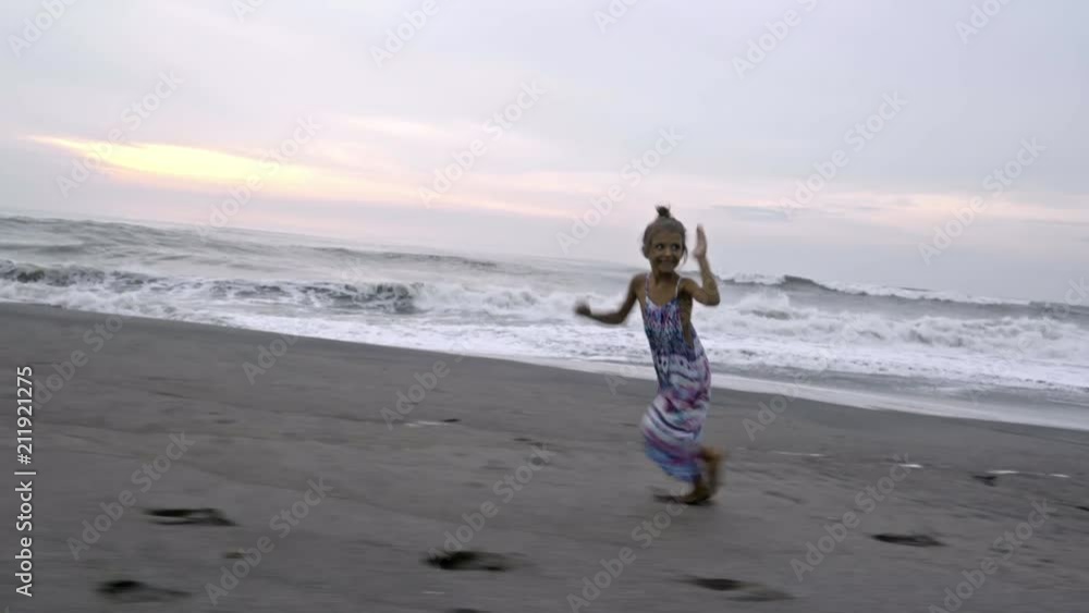 Cute little girl in summer dress smiling and showing thumbs up while running barefoot along ocean beach at sunrise Cute little girl in summer dress smiling and showing thumbs up while running barefoot along ocean beach at sunrise
