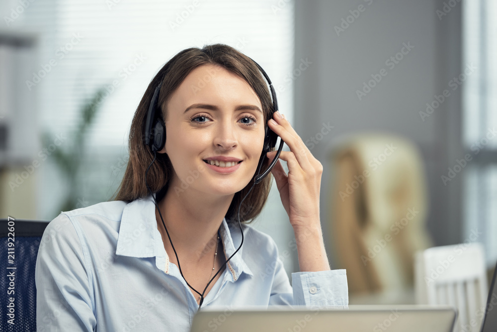 Young beautiful brunette working in call center as agent and communicating with customers wearing headset and smiling at camera Young beautiful brunette working in call center as agent and communicating with customers wearing headset and smiling at camera