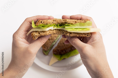 Bitten cheese and lettuce sandwich in hand, whole sandwich on white plate. Lunch, break, healthy snack concept