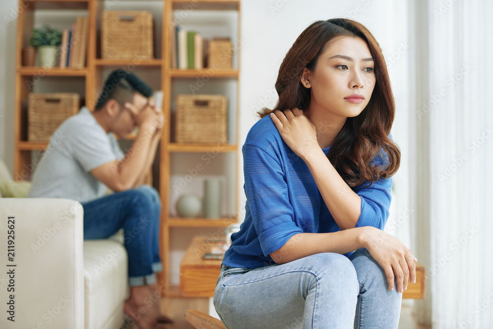 Sad Asian woman sitting alone with man on background being in conflict at home