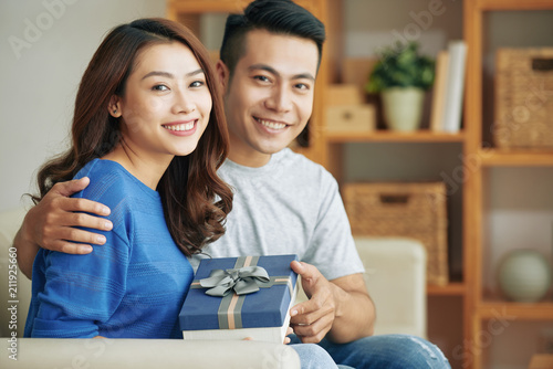 Wonderful happy Asian woman and man embracing on couch and holding decorated gift box smiling at camera