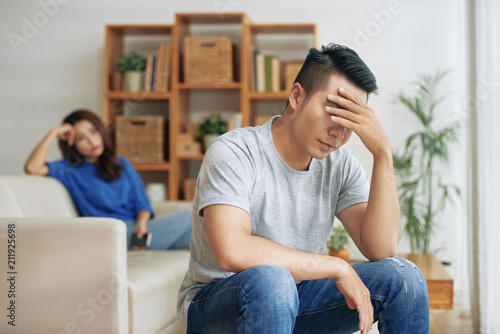 Upset Asian man sitting in living room with woman on background and covering eyes in depression having quarrel