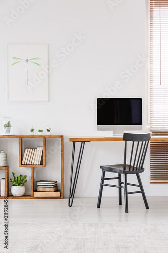 Real photo of a desk with a computer screen and a chair standing next to a wooden shelf in a simple office with blinds and a poster