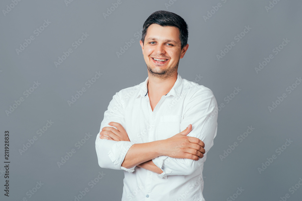 Handsome young man in white shirt keeping arms crossed smiling while standing against grey isolated background. People Emotions Lifestyle Fashion Business concepts Handsome young man in white shirt keeping arms crossed smiling while standing against grey isolated background. People Emotions Lifestyle Fashion Business concepts