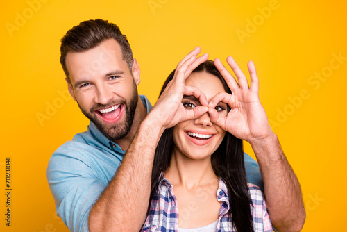 Ha-ha! Portrait of cool crazy couple, handsome man making binoculars with fingers to lover isolated on bright yellow background. Entertainment concept