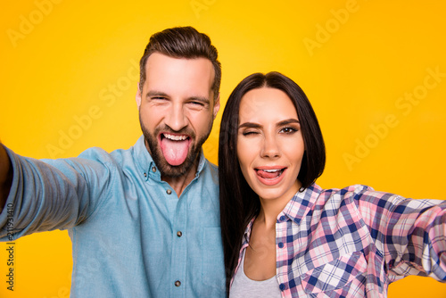 Self portrait of funny comic couple shooting selfie on front camera gesturing tongue out blinking with one eye isolated on vivid yellow background. Rest relax leisure concept