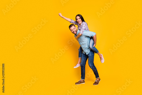 Full size portrait of playful carefree couple in piggy back style making super man sign with raised fist isolated on bright yellow background