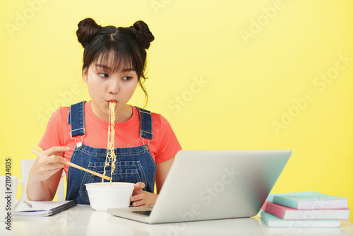 Asian woman sitting at the table with laptop and eating noodles
